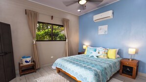 A bedroom with a bed, bedside tables, an air conditioner, a window with curtains, and a small wooden stool with a bucket. - Ocean Sounds Private Apt for couples! Marina view (QUEPOS)