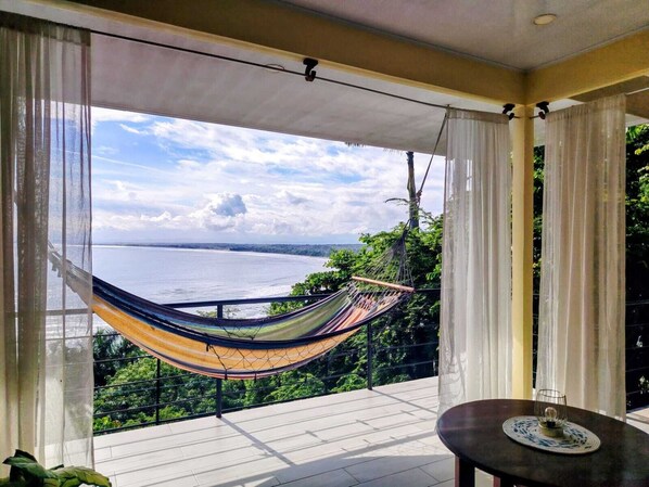 A balcony with a hammock, a table, and a view of a body of water.