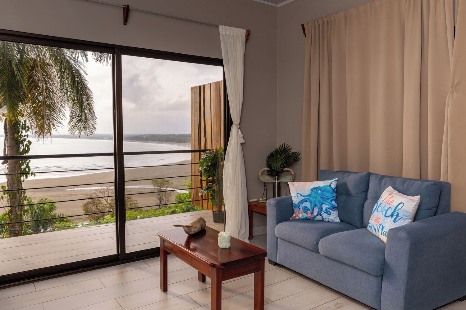 A room with a view of the beach, a blue sofa, a wooden table, and a potted plant.