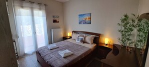A hotel room with a bed, bedside lamps, a radiator, a window with curtains, a wall-mounted picture, and a potted plant.