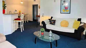 Een woonkamer met een glazen salontafel, een bank met gekleurde kussens, een eethoek met een witte tafel en stoelen, en een keuken met een spoelbak en aanrecht.
