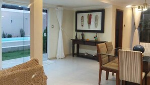 A spacious living area with a glass coffee table, a woven pouf, and a dining table with chairs. A framed artwork is displayed on the wall, and a pool is visible through the glass door. - Recanto da Sulla, Seu Refúgio Perfeito: Piscina Privativa, Área Gourmet e Charme (Jauá Camaçari)