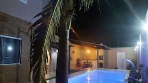 A swimming pool with a tiled deck, surrounded by a building with a modern design, including a visible kitchen area and a dining space. - Recanto da Sulla, Seu Refúgio Perfeito: Piscina Privativa, Área Gourmet e Charme (Jauá Camaçari)