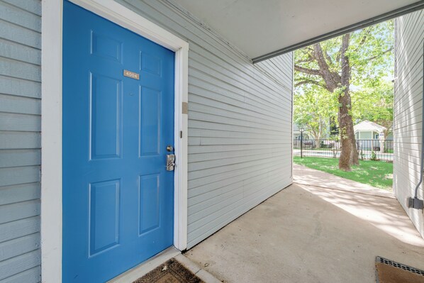 A blue door with a number plate, a concrete porch, and a view of a tree and a building in the background.