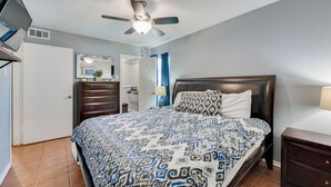 Quarto com cama, ventoinha no teto, cômoda, espelho e TV sobre um suporte.