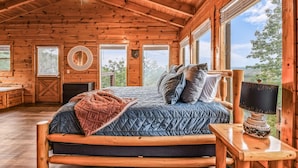 Un dormitorio de cabaña de madera acogedor, con una cama, ventilador de techo y ventanas amplias que ofrecen vistas al exterior.