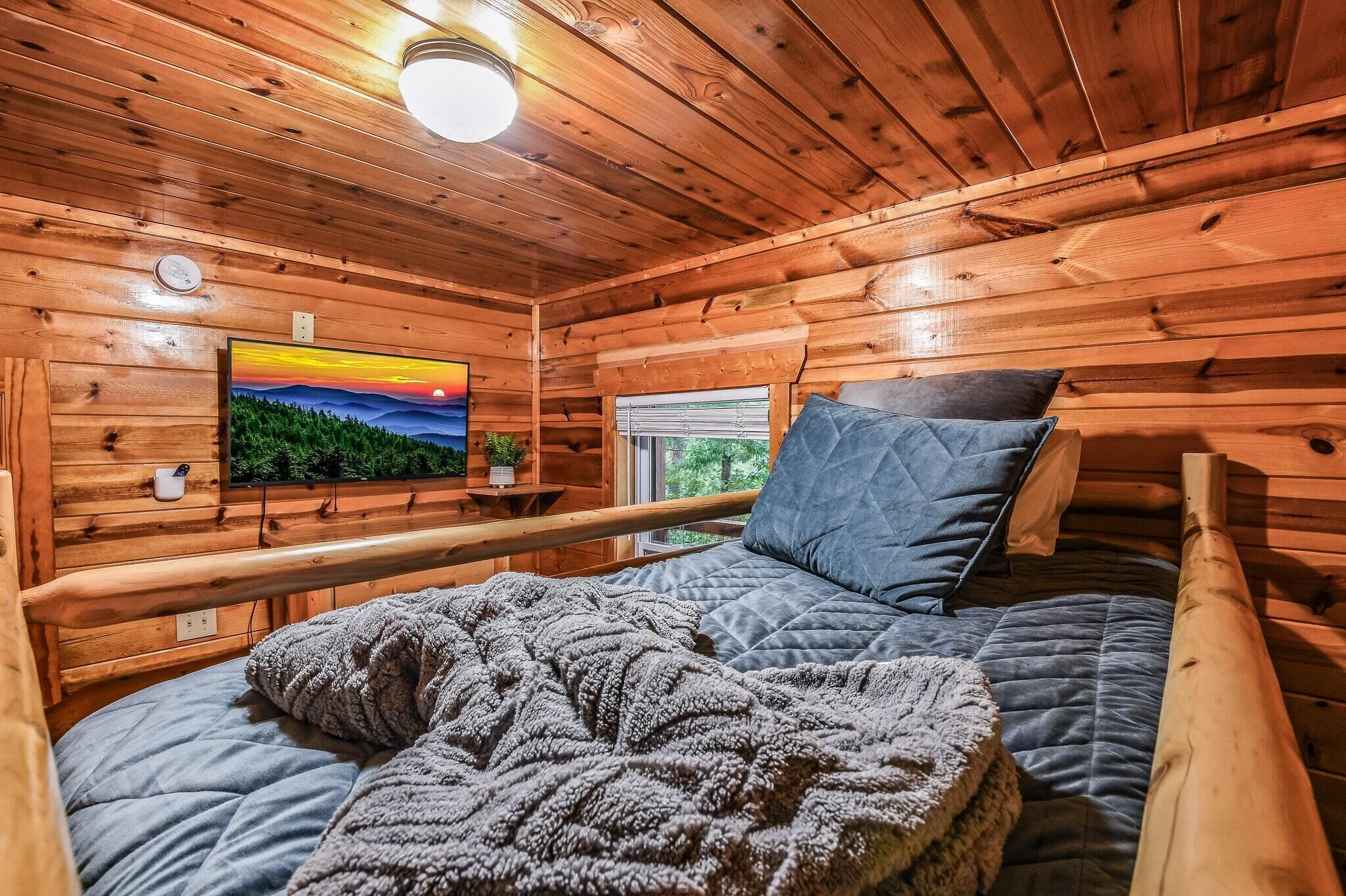 A cozy cabin room with a bed, a mounted TV, a window, and wooden walls.