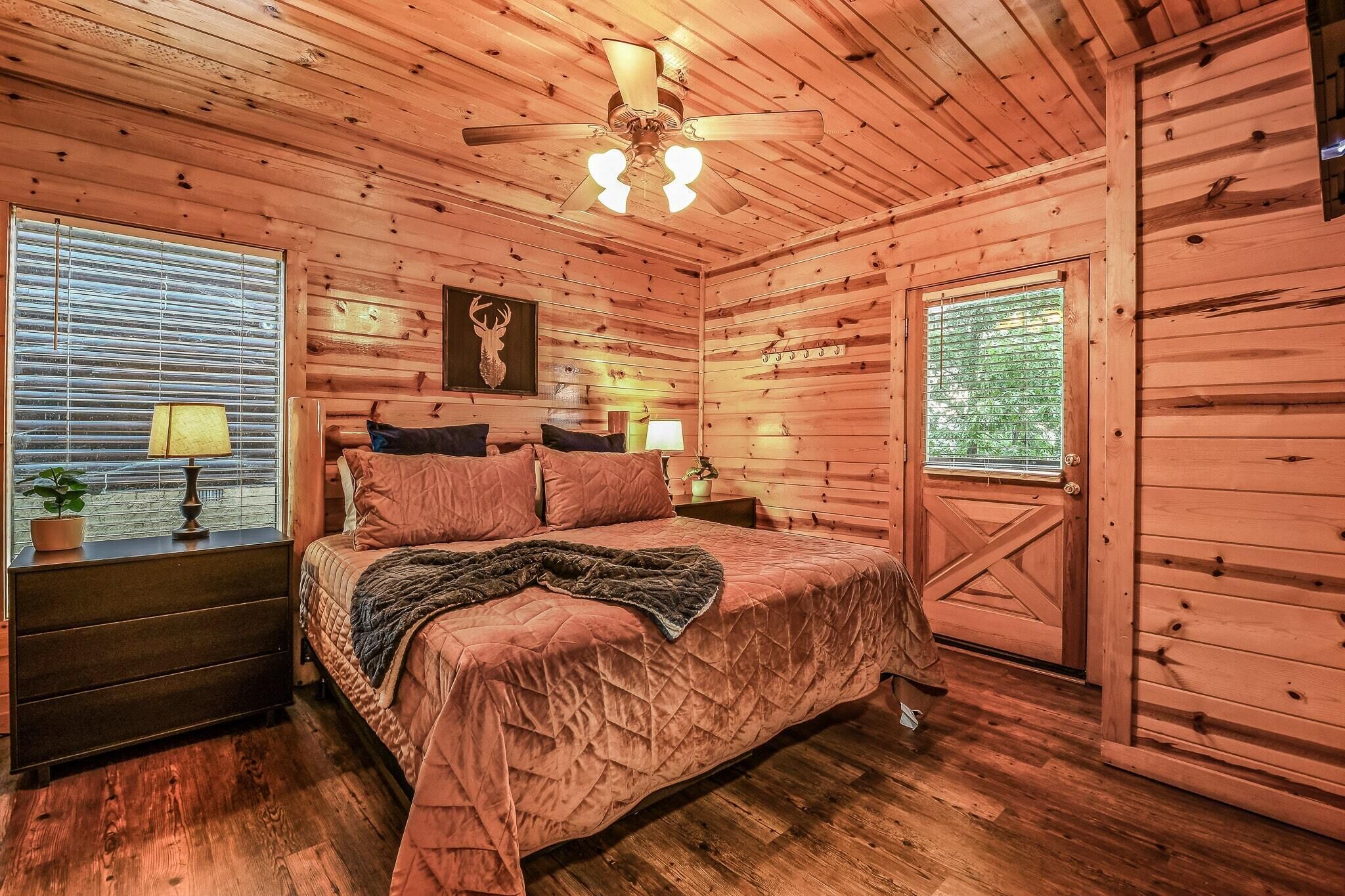A wooden cabin bedroom with a bed, two bedside tables, a ceiling fan, and a window with blinds.