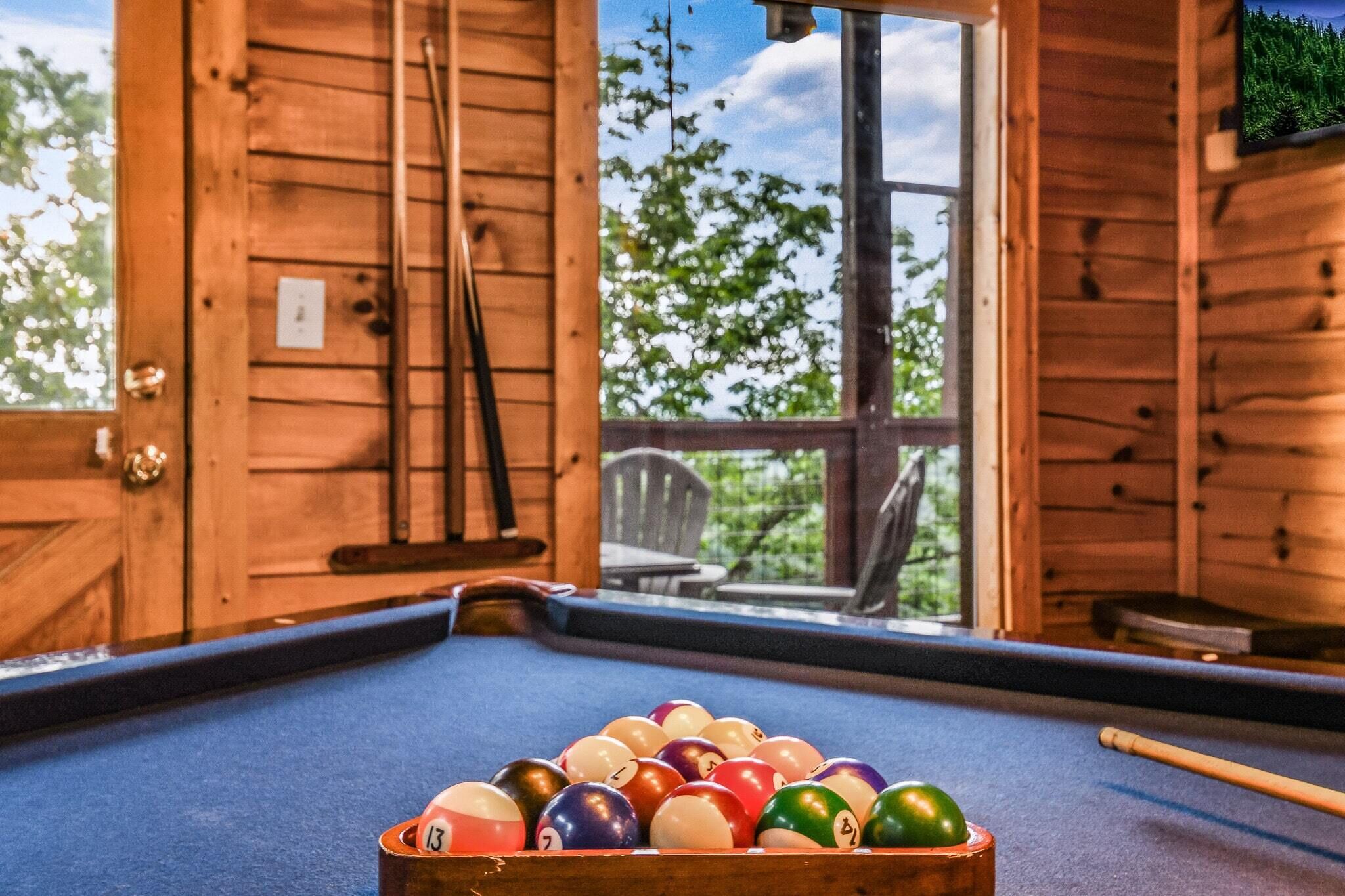 Une table de billard avec des billes, dans une pièce en bois dotée de grandes fenêtres.