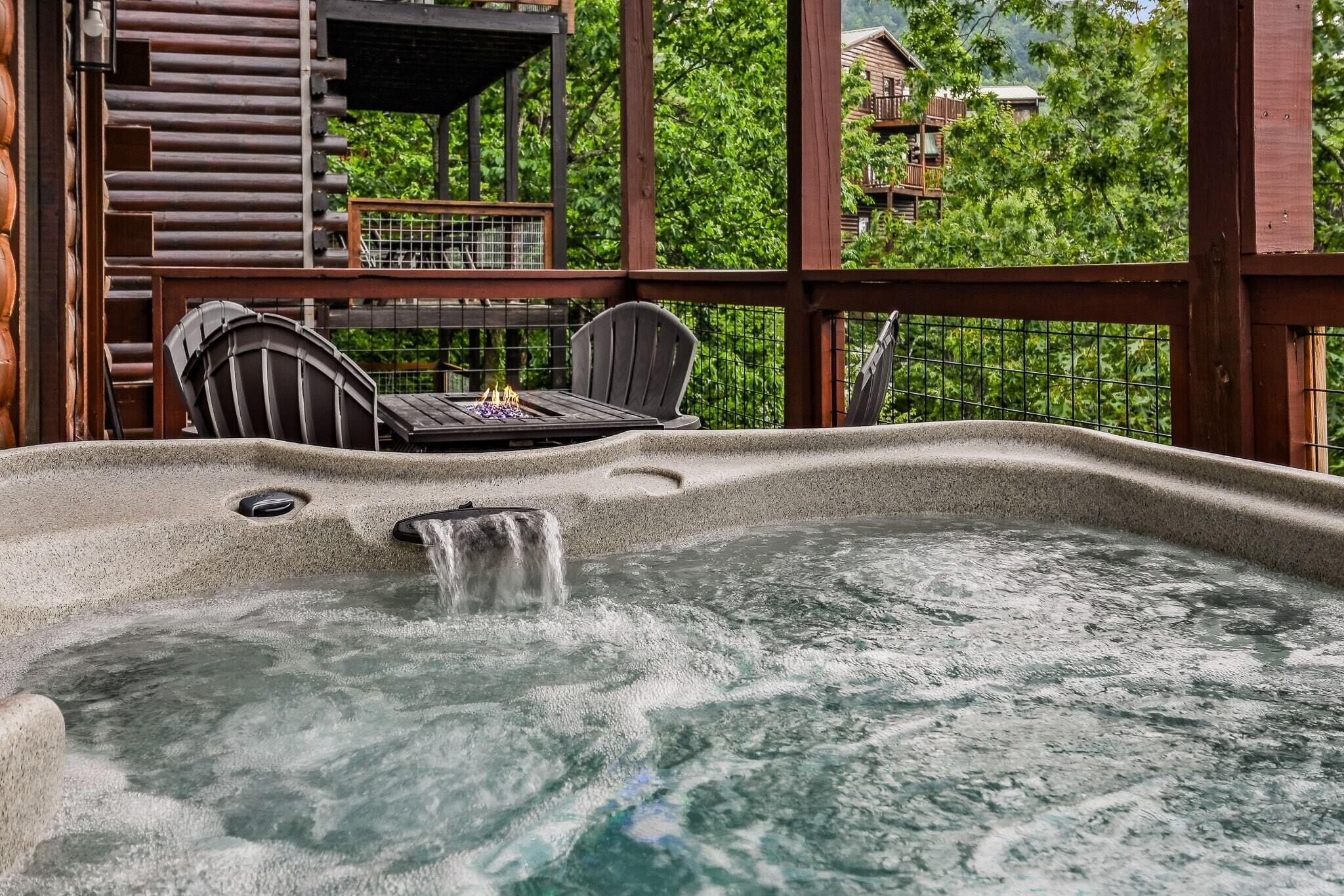 A hot tub with a waterfall feature, surrounded by wooden deck furniture and a view of a forested area.