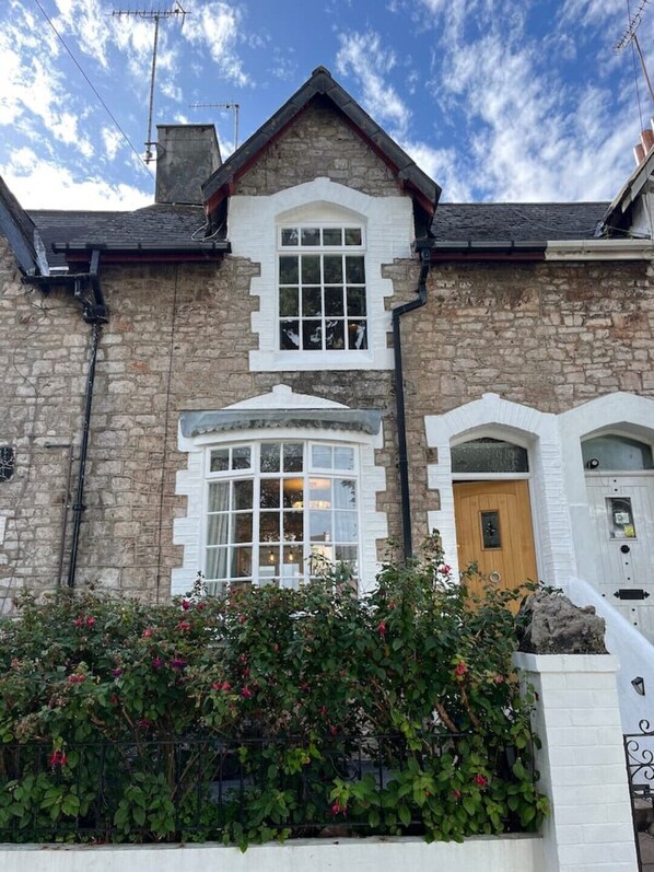 A two-story stone house with a prominent bay window, a wooden front door, and a small porch area. - Charming Central Torquay Cottage (Torquay)