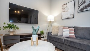 Una sala de estar moderna con un sofá gris, una mesa de centro redonda blanca, un televisor de pantalla plana montado en la pared y una lámpara de pie.
