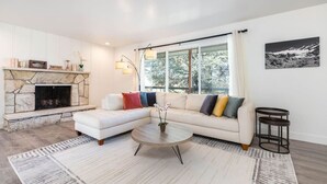 Un salon moderne avec une cheminée en pierre, un canapé d’angle agrémenté de coussins colorés, une table basse ronde et une grande fenêtre donnant sur la forêt.