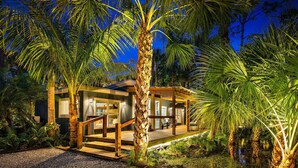 Een modern huis met een houten terras, omgeven door palmbomen en een kleine vijver.