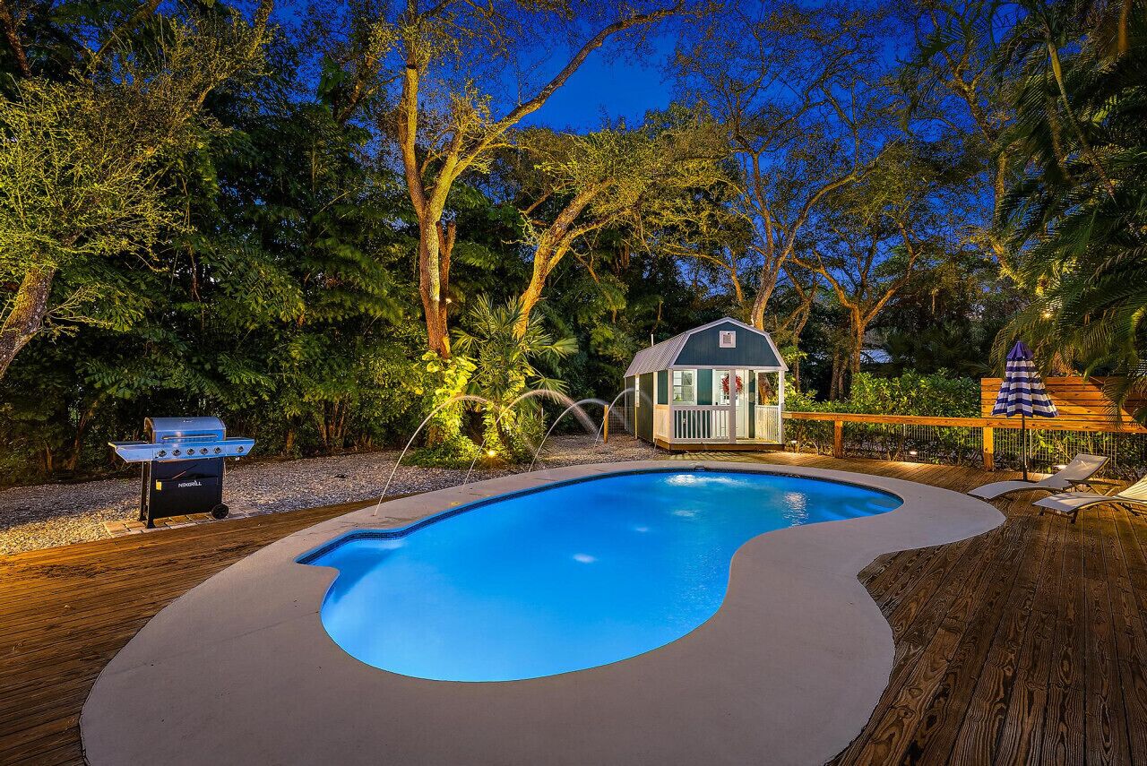 Une piscine avec une terrasse en bois, un barbecue et une petite maison dotée d’un porche.