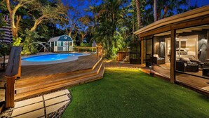 Een zwembad met houten terras, een klein huisje en een loungeruimte met glazen wanden.