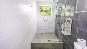 A bathroom with white tiled walls, a shower with a glass door, a window with greenery, and a toilet with a paper towel holder. - Cosy, personal, family-friendly nature retreat surrounded by trees and fresh air (Aburi)