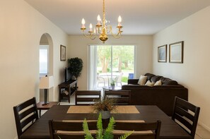 A dining area with a wooden table, chairs, a chandelier, and a view of the outdoors through large windows.