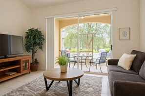 A living room with a TV, a sofa, a coffee table, and a sliding glass door leading to an outdoor patio with chairs and a table.
