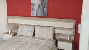 Un dormitorio con pared roja, cabecero de madera, cama con ropa de cama a rayas, dos mesitas de noche con lámparas y un cuadro enmarcado en la pared.