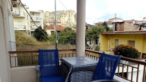 Un balcón con una mesa a cuadros y sillas azules, con vista a una zona residencial que incluye edificios y una montaña al fondo.