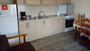 Cocina compacta con armarios de madera clara, un refrigerador blanco, cafetera y un lavamanos. Se ve una silla de madera con un cojín estampado y una mesa con mantel rojo y blanco.