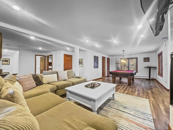 A spacious living room with a beige sectional sofa, a white coffee table, and a pool table.