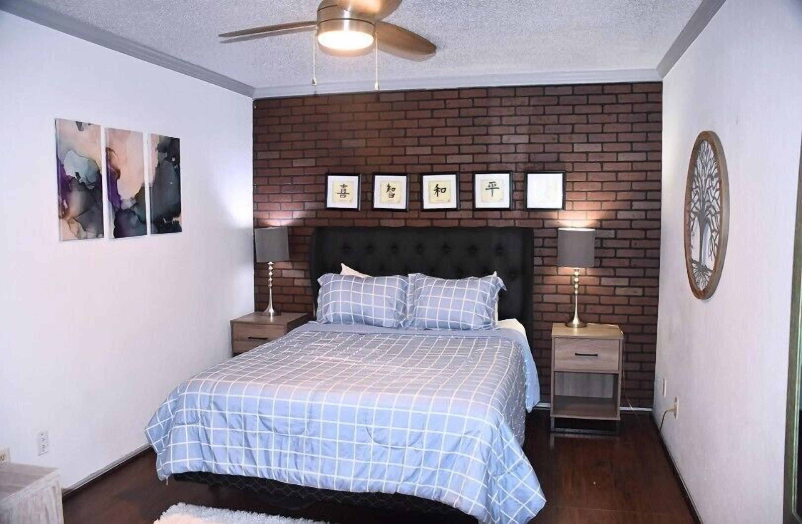 A bedroom with a bed, two bedside tables, a ceiling fan, and a brick wall.