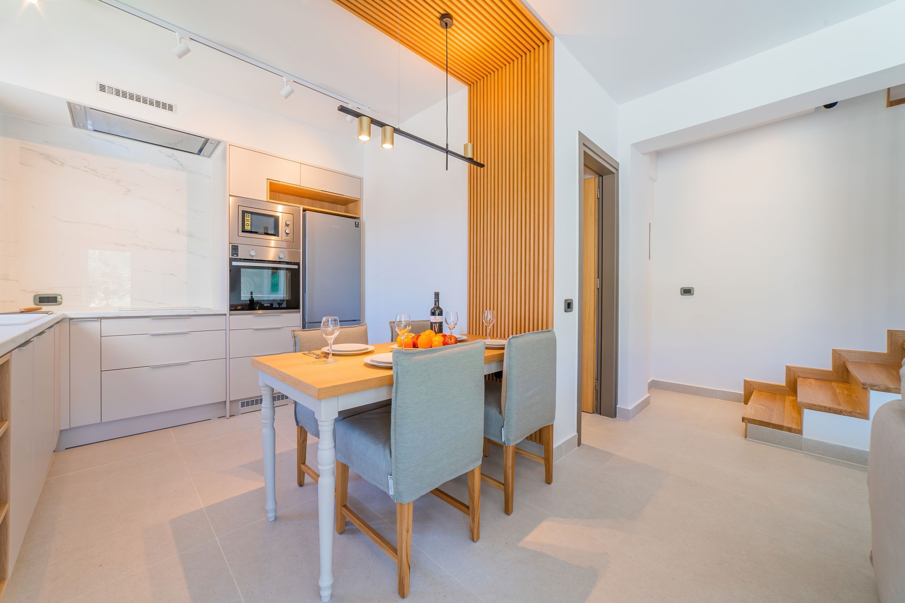 Una cocina moderna con electrodomésticos integrados, un comedor con mesa y sillas de madera, y una escalera que sube a otro nivel.