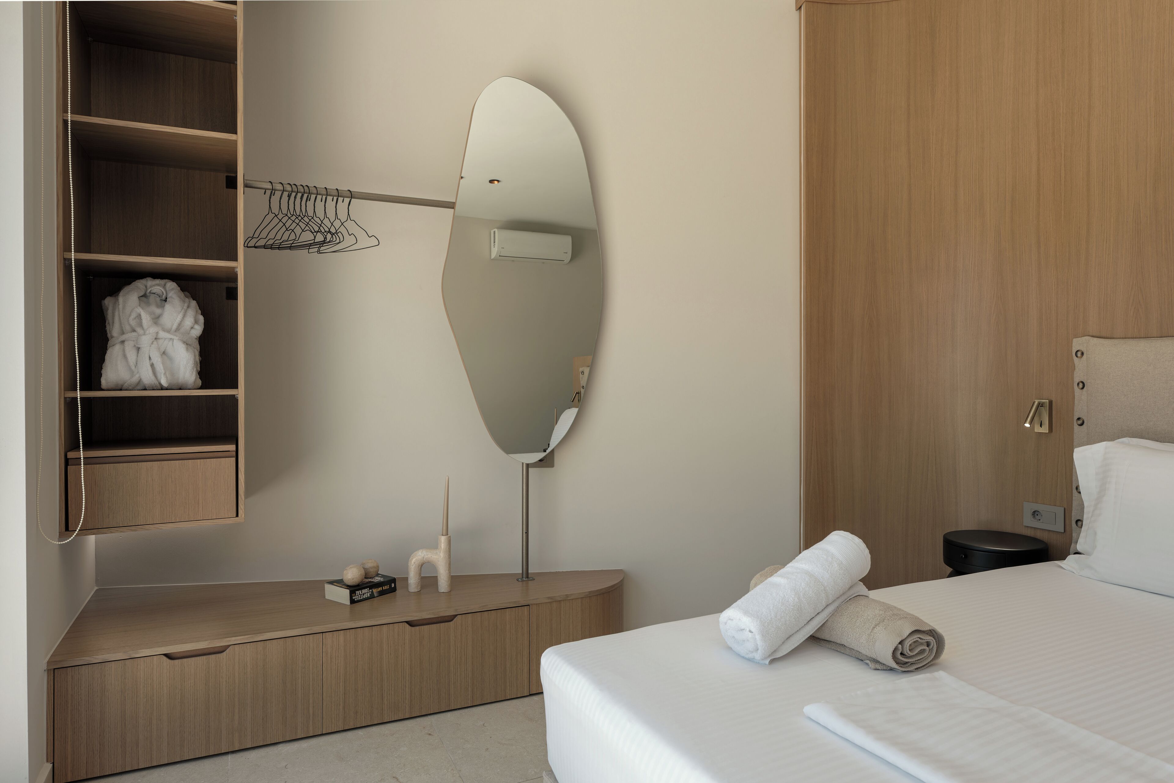 Una habitación de hotel con un armario de madera, una mesita de noche con lámpara, un espejo y una cama bien hecha con una toalla enrollada.