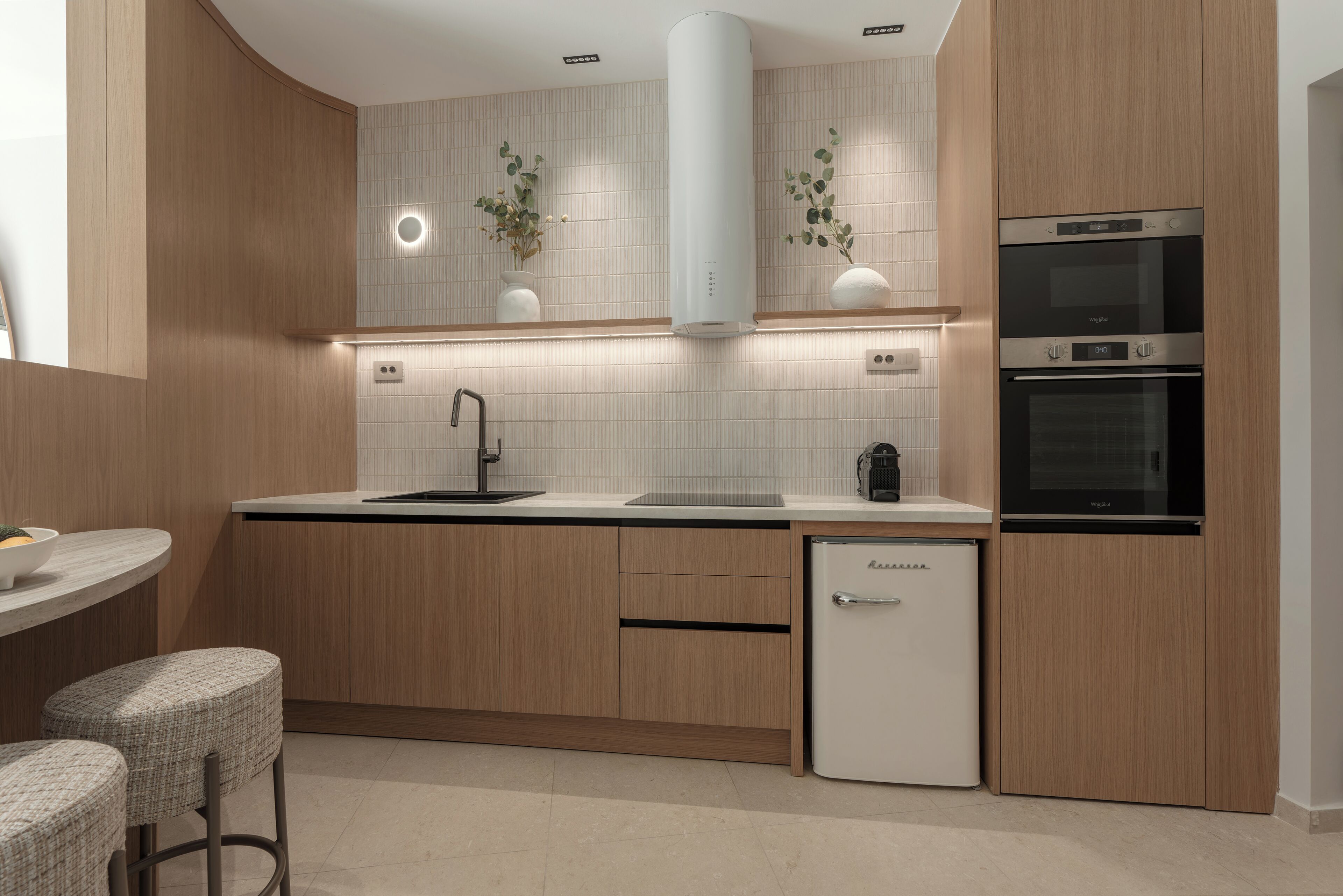 Una cocina moderna con armarios de madera, horno empotrado, refrigerador y una zona de bar con taburetes.