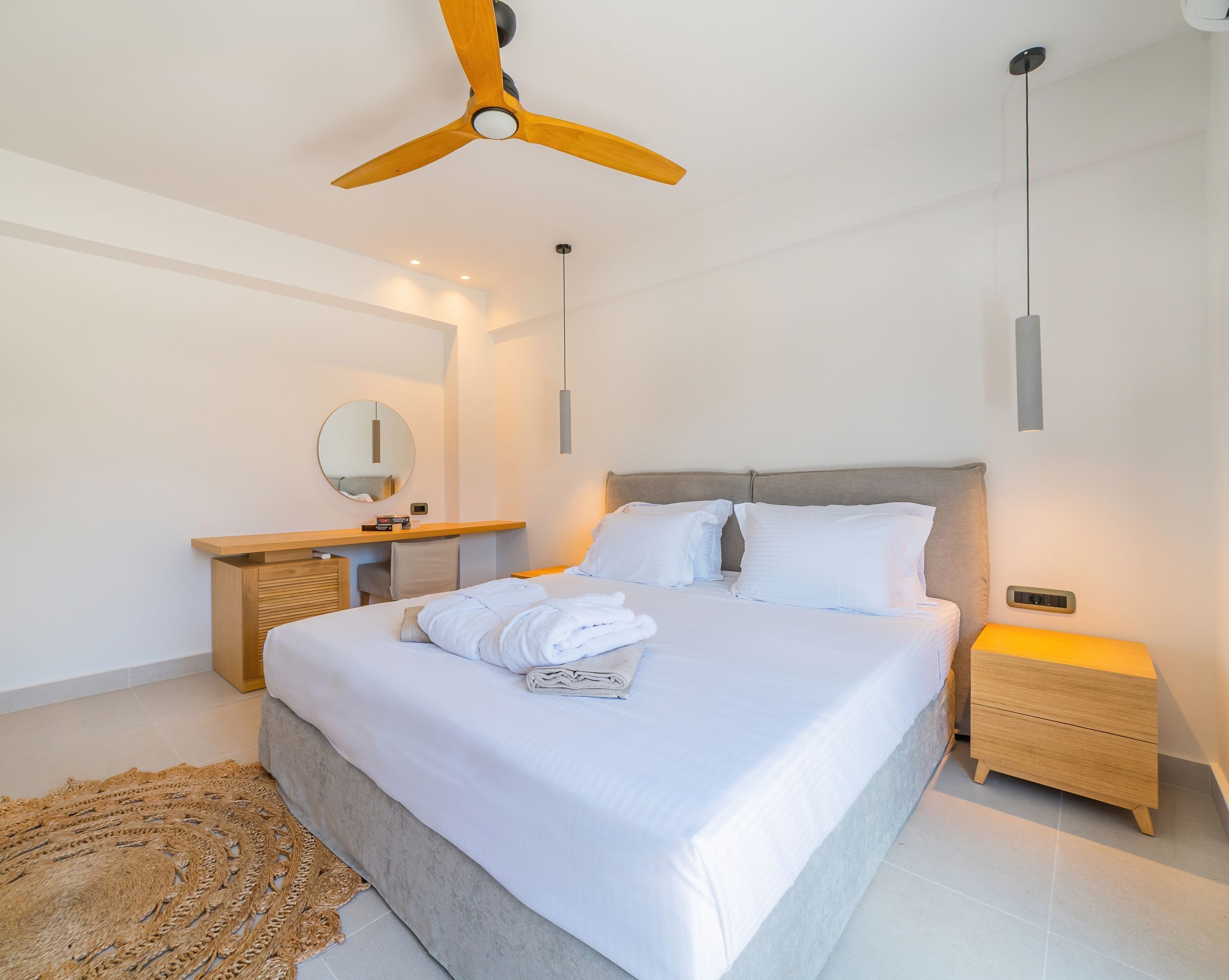 Un dormitorio moderno con una cama grande, cabecera de madera, mesitas de noche, un ventilador de techo y un espejo redondo.