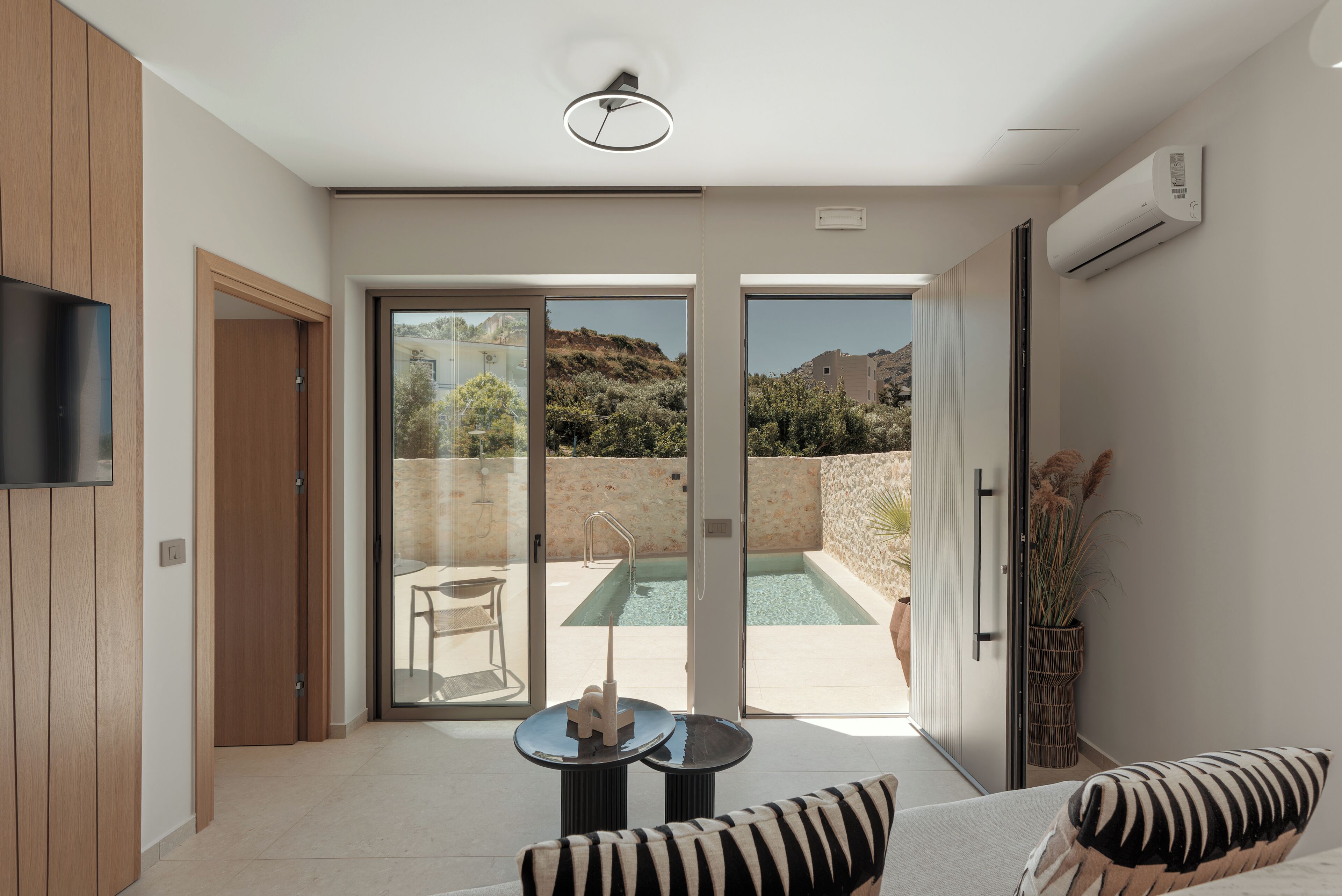 A modern interior with a sliding glass door leading to an outdoor pool area, a flat-screen TV mounted on the wall, and a small round table with a chair.