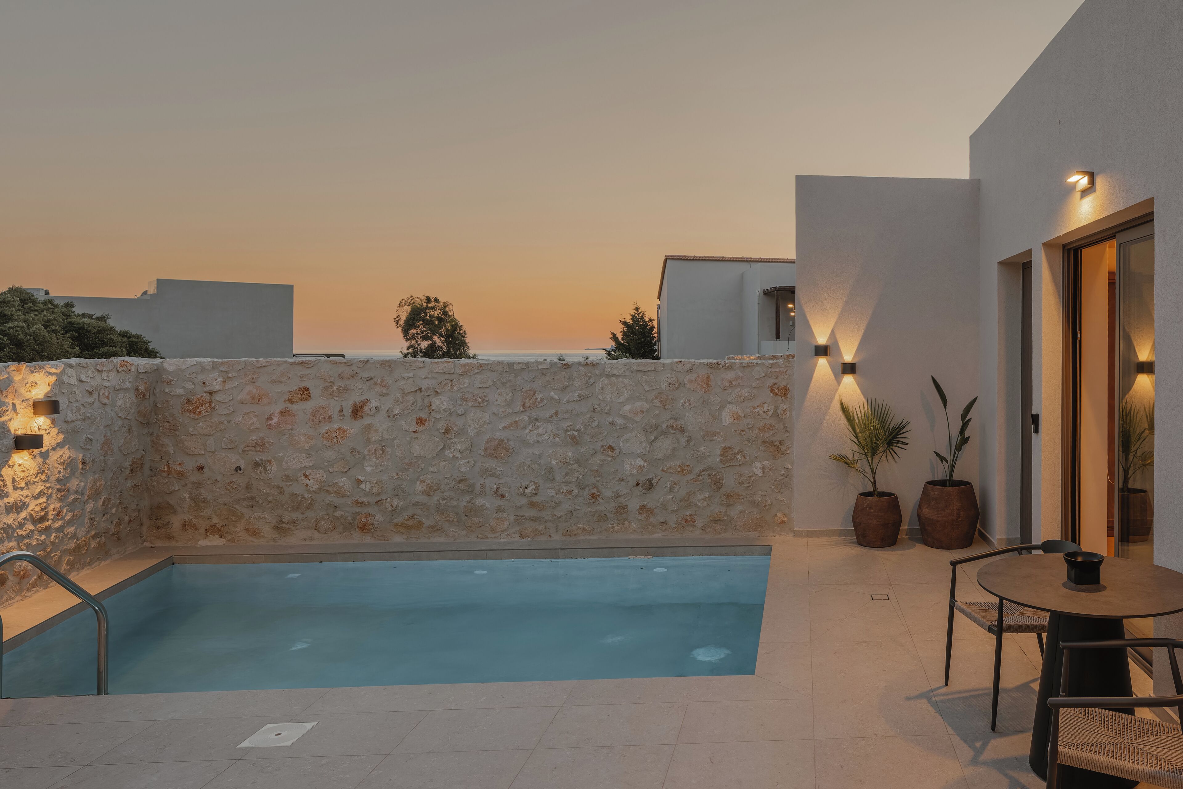 A rooftop pool with stone walls, potted plants, and a table with chairs.