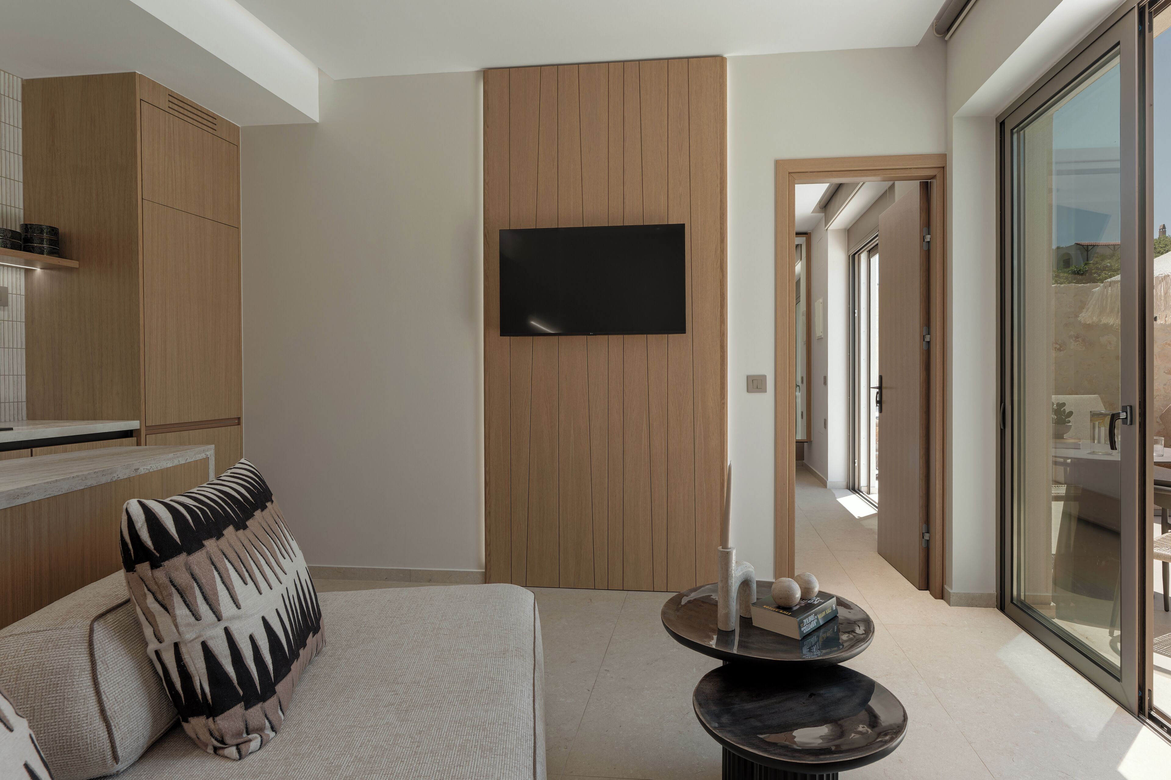 Een moderne woonkamer met een bank, een flatscreen-tv bevestigd aan een houten paneelwand, een kleine ronde tafel met decoratieve objecten en een glazen deur die leidt naar een buitengebied.