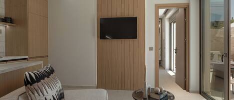 Een moderne woonkamer met een bank, een flatscreen-tv bevestigd aan een houten paneelwand, een kleine ronde tafel met decoratieve objecten en een glazen deur die leidt naar een buitengebied.