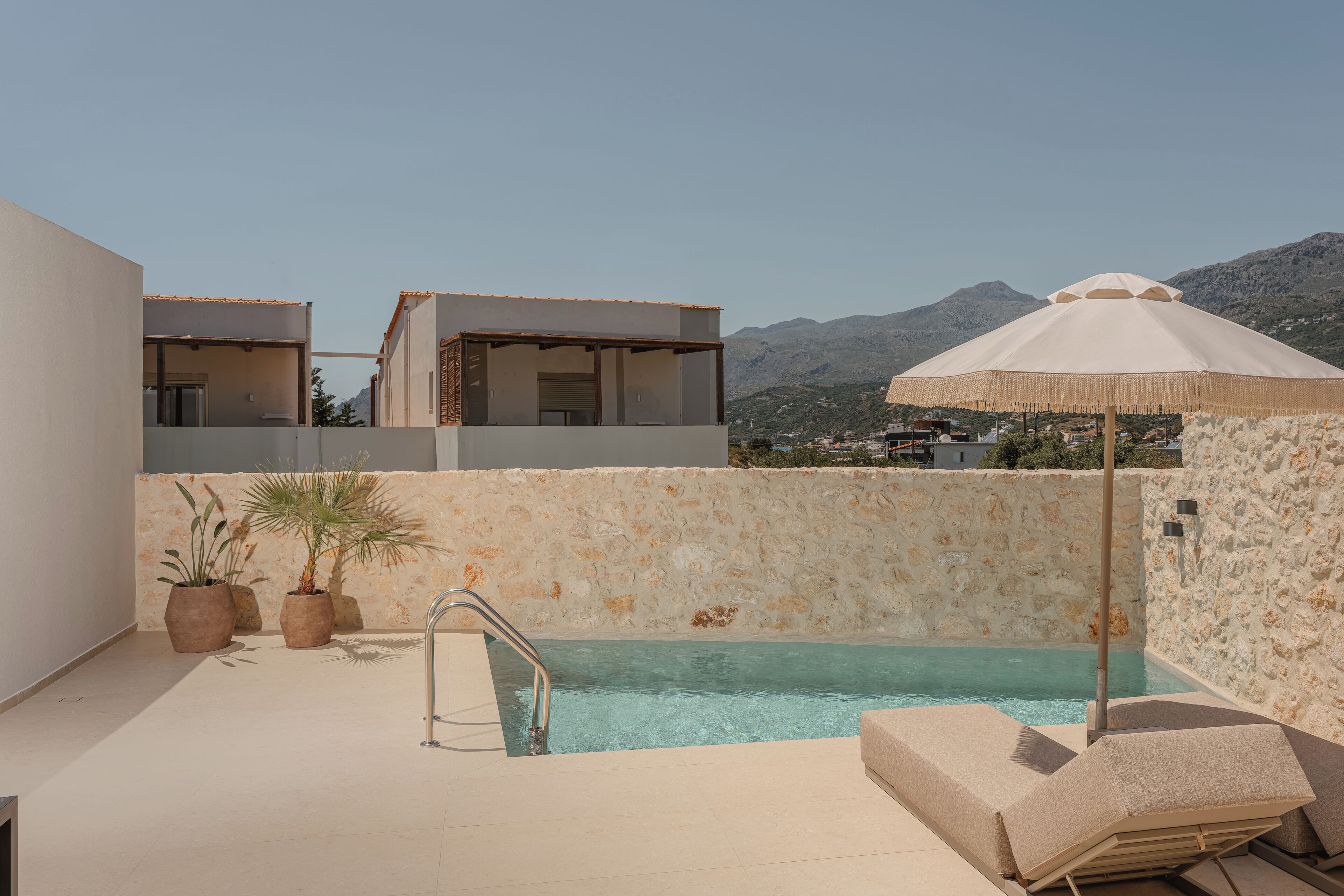 A rooftop pool with a stone wall, lounge chairs, and potted plants.
