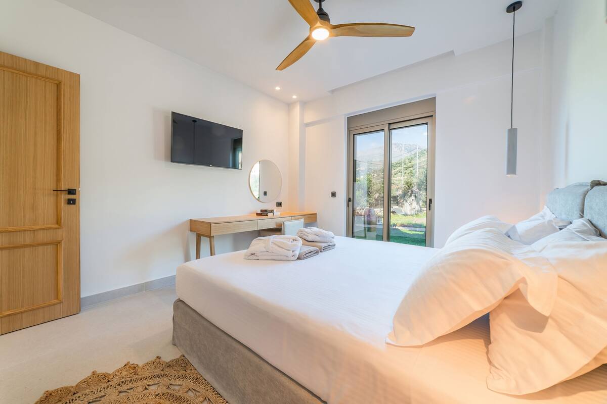 A modern bedroom with a large bed, a wooden door, a desk with a flat-screen TV, a ceiling fan, and a view of greenery outside.