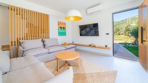 A modern living room with a sofa, a round wooden coffee table, and a large window offering a view of greenery. - Increteblue Suites (Agios Vasileios)