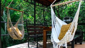 Duas redes, uma com padrão de folhagem natural e outra com padrão em branco e amarelo, penduradas em uma estrutura de madeira em um ambiente verdejante e arborizado.