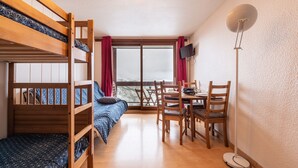 Quarto com beliche, mesa de jantar com cadeiras, televisão fixada na parede e uma grande janela com cortinas.