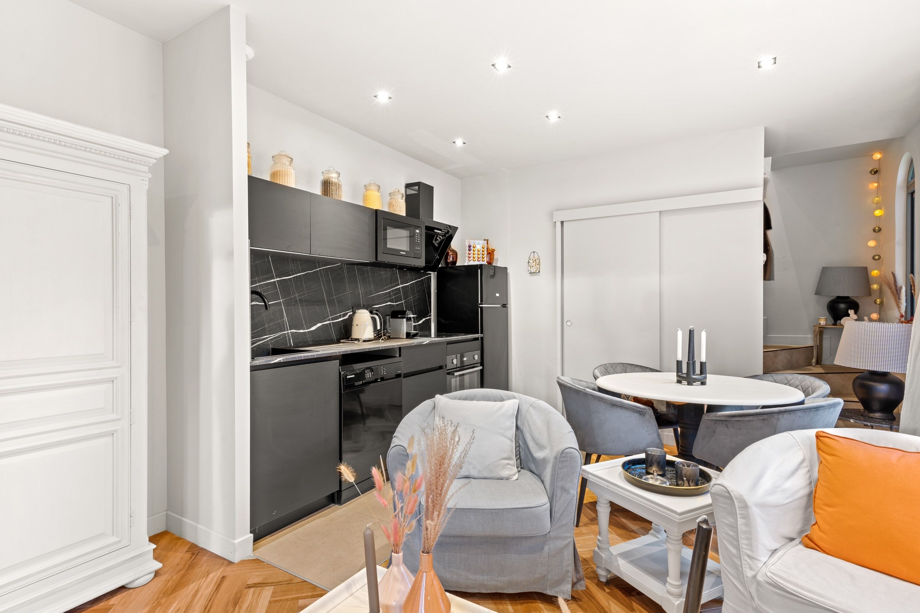 A modern kitchen with dark cabinetry, a black marble backsplash, and a white countertop. There is a dining area with a round table and grey chairs.