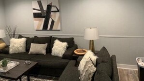 Ein modernes Wohnzimmer mit einem grauen Sofa, weißen, flauschigen Kissen, einem Couchtisch mit dekorativen Accessoires und einer Stehlampe.