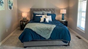 Un dormitorio con cabecera capitonada, una cama grande con ropa de cama azul, dos mesitas de noche con lámparas y obras de arte en las paredes.