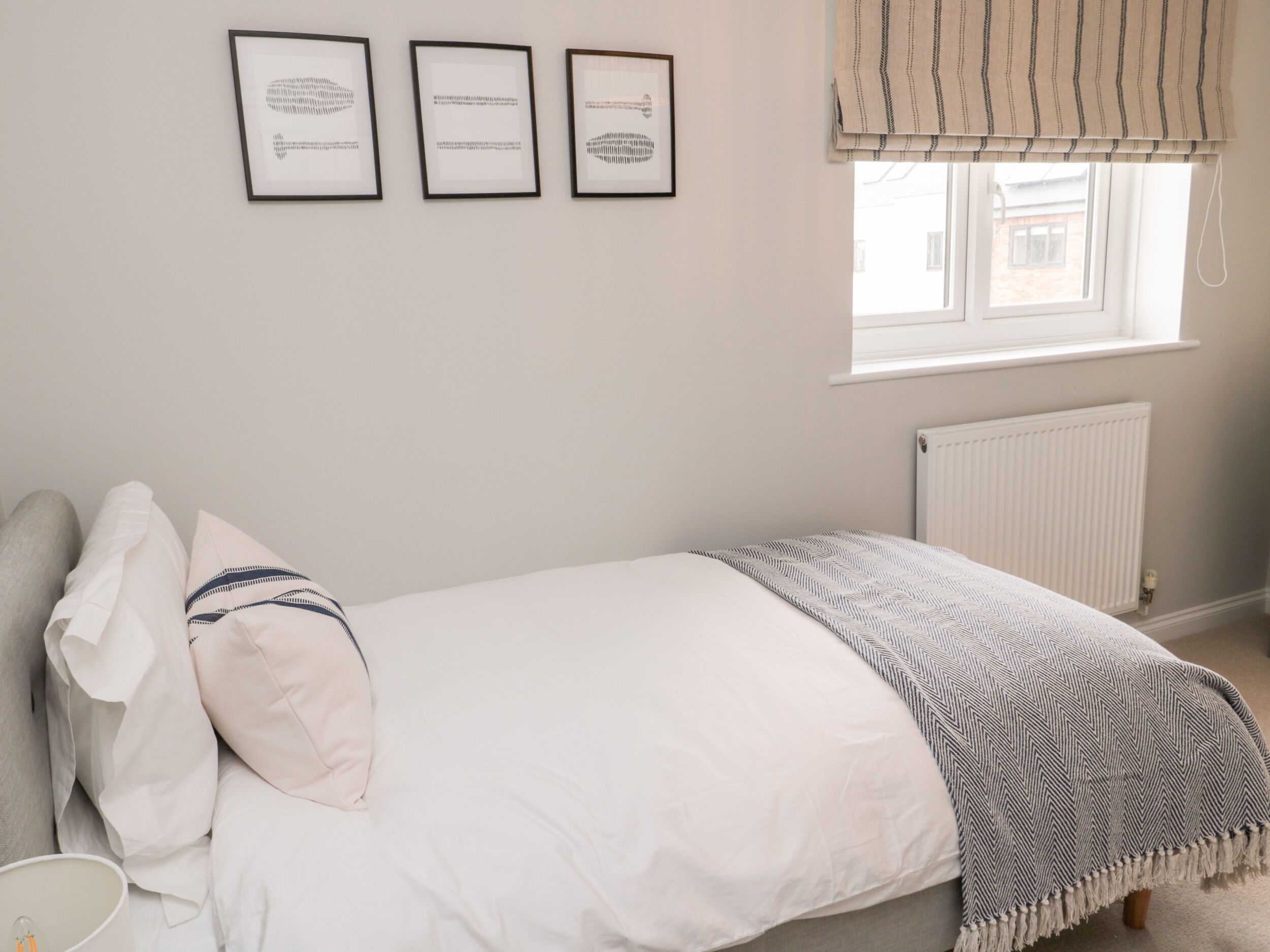 Une chambre à coucher avec un lit, des oreillers, une couverture, une fenêtre avec des stores et trois tableaux encadrés accrochés au mur.