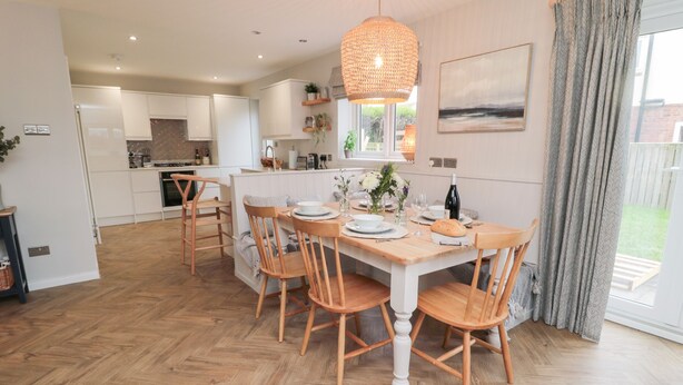 A modern kitchen with white cabinets, a dining area with wooden chairs, a hanging light fixture, and a view of the outdoors through a window. - Driftwood Sands (Chathill)