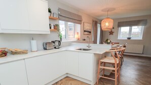 A modern kitchen with white cabinets, a wooden herringbone floor, a built-in island with a sink, and a dining area with wooden chairs. - Driftwood Sands (Chathill)