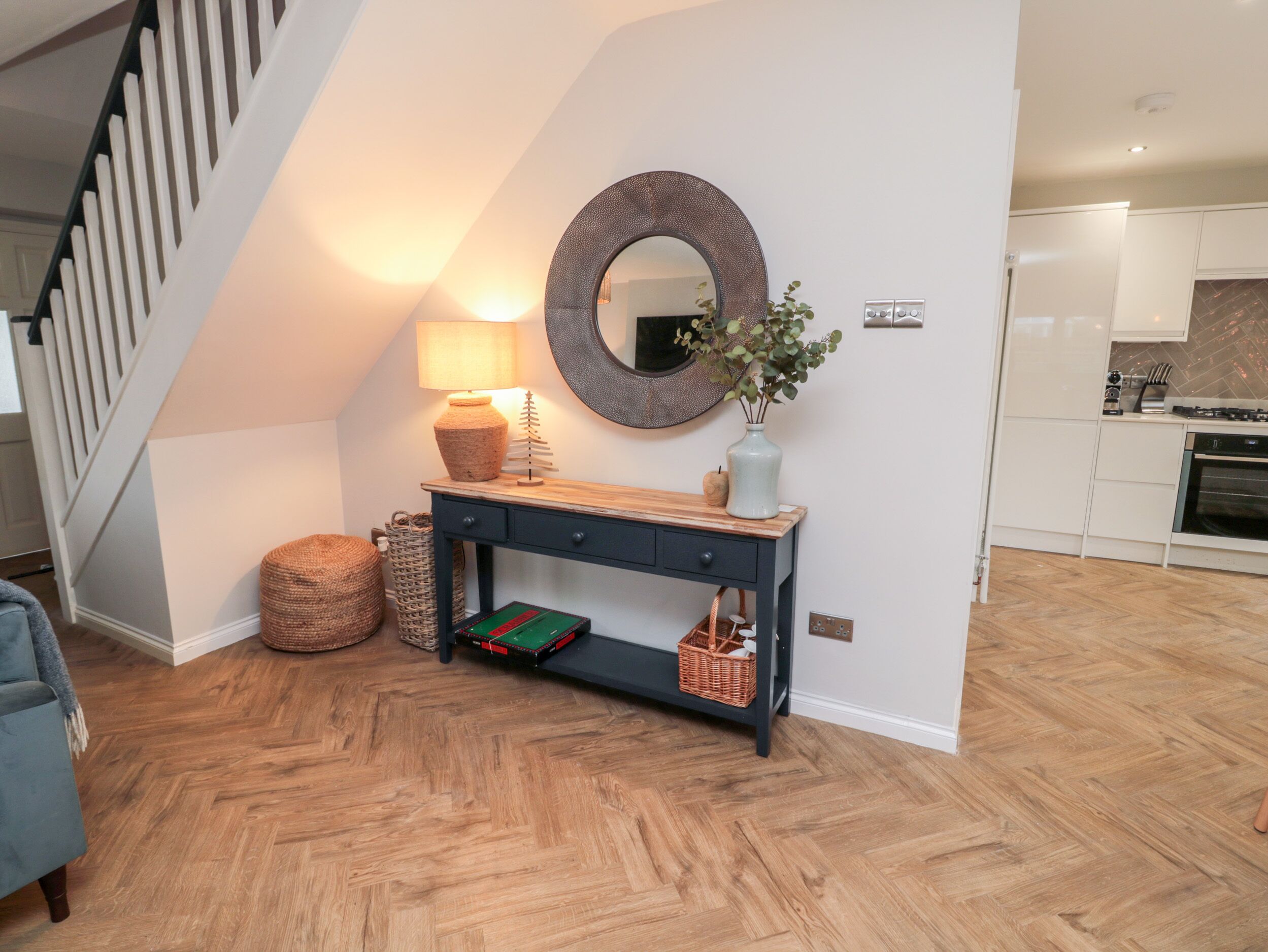 A modern living room with a herringbone wood floor, a dark blue console table with a lamp, a mirror, and a vase with greenery.