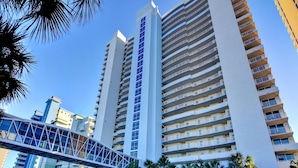 A high-rise hotel with multiple floors, a pedestrian bridge, and palm trees. - Majestic Beach Towers I by Southern Vacation Rentals (Panama City Beach)
