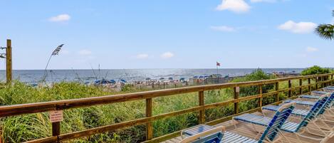 Una terraza de madera con sillas azules, barandilla y vista a la playa con sombrillas y el océano.